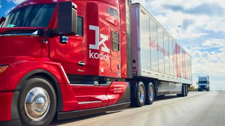 Red Kodiak self-driving semi-truck on a highway with a silver self-driving trailer under a partly cloudy sky.