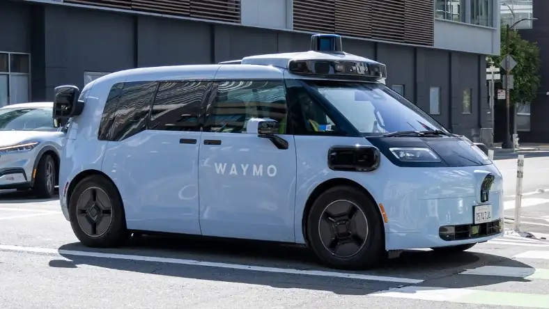 White Waymo driverless vehicle with rooftop sensor array at a city street crossing alongside other traffic