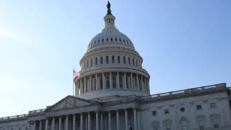 The dome and columned facade of the US Capitol building with an American flag flying against a clear blue sky